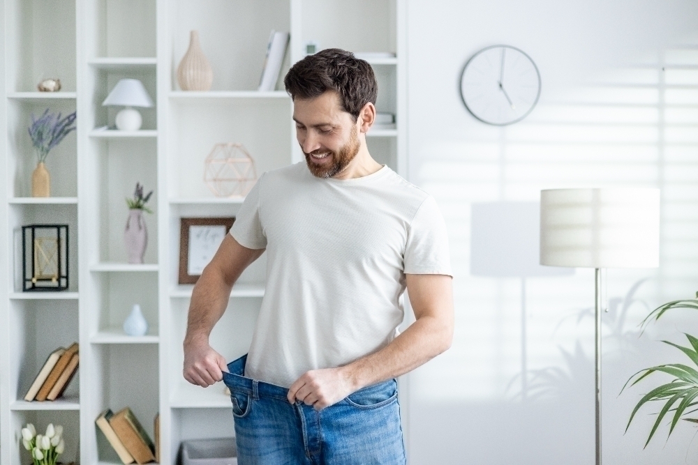 A joyful man showcasing his weight loss by pulling his loose jeans at the waist in a bright modern